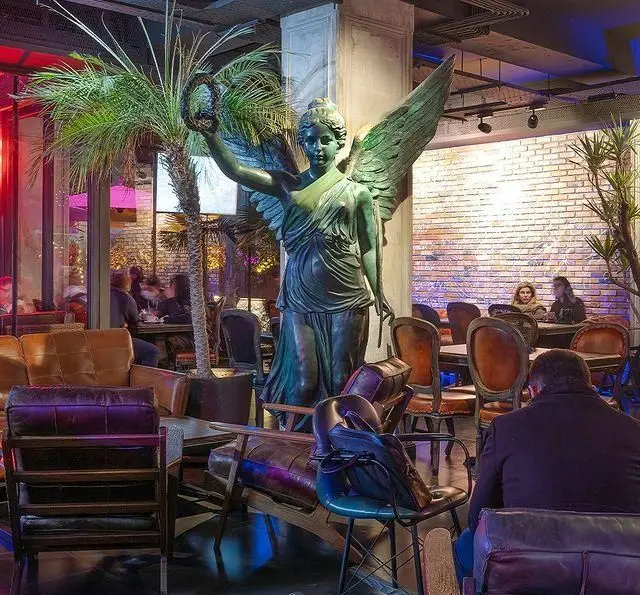 فضای داخلی کافه رستوران مونلانژ با دکوراسیون لوکس، مجسمه زن بالدار برنزی، مبلمان چرمی و درخت نخل در کنار مشتریان.