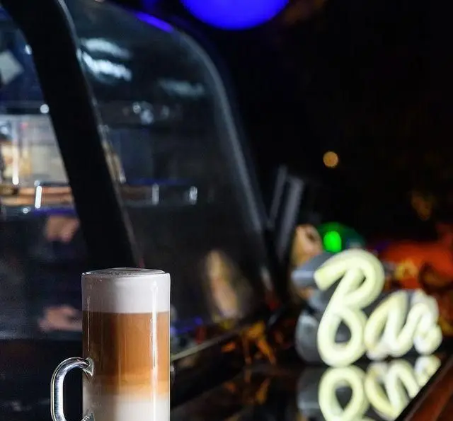 یک لیوان لاته ماکیاتو با لایه‌های قهوه و شیر، آماده سرو در فضای شبانه و دنج کافه رستوران مونلانژ. لذت نوشیدنی گرم.