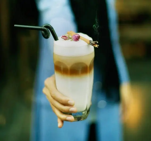 لاته لایه‌لایه جذاب با نی مشکی، تزیین رزهای خشک و سیگار روشن روی فوم در کافه رستوران مونلانژ.