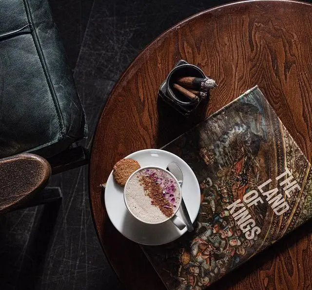 کافه رستوران مونلانژ: فضایی دنج و لوکس با لاته گرم مزین به گلبرگ رز، بیسکویت، کتاب "The Land of Kings" و سیگار برگ روی میز چوبی.