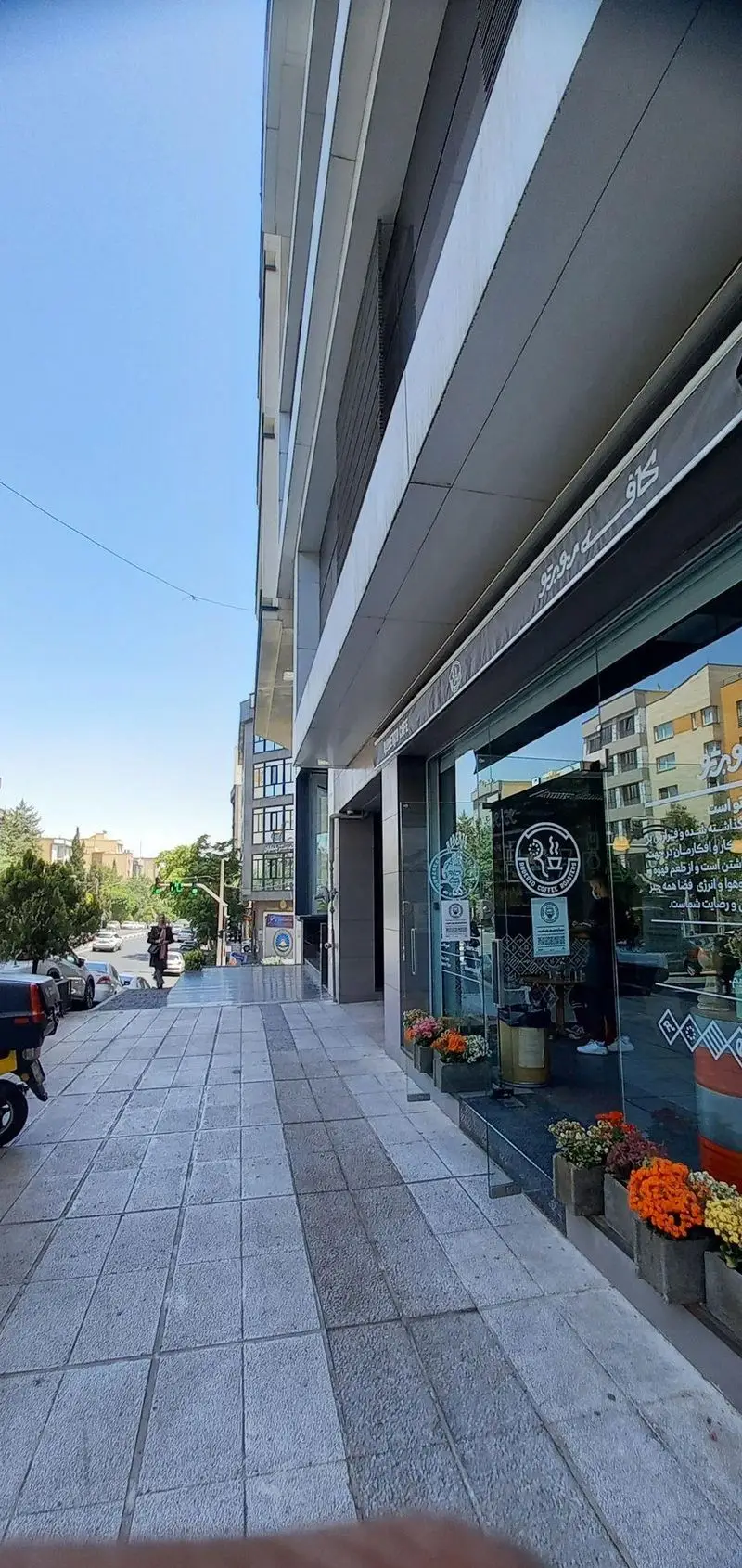 کافه روبرتو، نمایی زیبا از فضای بیرونی و ویترین شیشه‌ای با لوگوی ROBERTO COFFEE ROASTERS و گل‌های رنگی در پیاده‌رو شهری.