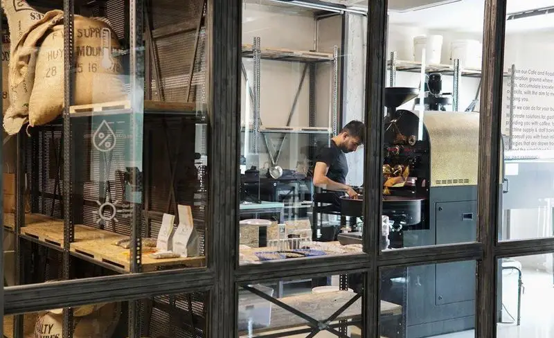 کافه روبرتو: مردی در حال رست قهوه با دستگاه پیشرفته در کارگاه. دانه های سبز قهوه و تجهیزات آماده سازی در محیطی صنعتی و مدرن.