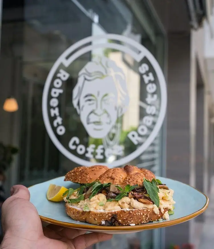 ساندویچ تازه و خوش‌طعم در کافه روبرتو، حاوی قارچ و سبزیجات معطر، با نان کنجدی، ایده‌آل برای صبحانه یا میان‌وعده. لوگوی Roberto Coffee Roasters در پس‌زمینه.