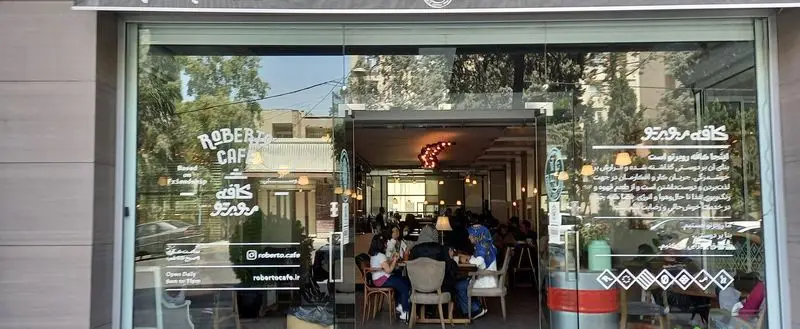 ورودی کافه روبرتو، یک کافی شاپ گرم و دوستانه، با شیشه های بزرگ، دکوراسیون داخلی جذاب، مشتریان و اطلاعات تماس و شعار 'بر پایه دوستی'
