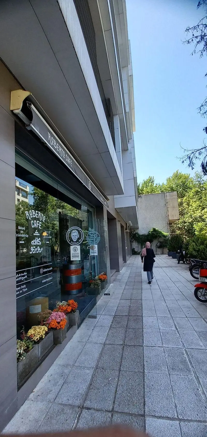 نمای بیرونی کافه روبرتو با تابلوی مشکی، ورودی شیشه‌ای، گلدان‌های رنگارنگ و پیاده‌رو. فضایی دلنشین برای مشتریان.