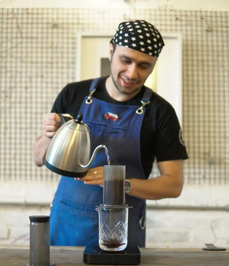مردی با کلاه ستاره‌دار و پیش‌بند آبی در حال دم کردن قهوه. باریستای ماهر کافه روبرتو با کتری مخصوص، قهوه خوش‌طعم و تازه برای شما آماده می‌کند.
