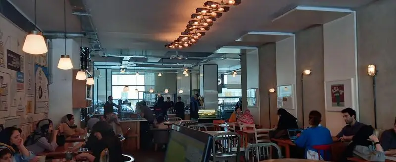 فضای داخلی مدرن و شلوغ کافه روبرتو، با نورپردازی خاص و مشتریانی که در حال کار، گفتگو و صرف غذا در محیطی دلپذیر هستند.