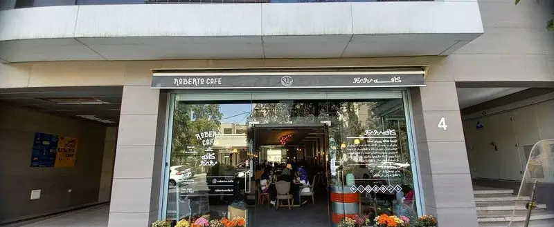 کافه روبرتو، نمای بیرونی جذاب با تابلو انگلیسی و فارسی، درب شیشه‌ای، دکوراسیون داخلی روشن و مشتریان در حال لذت بردن از قهوه.