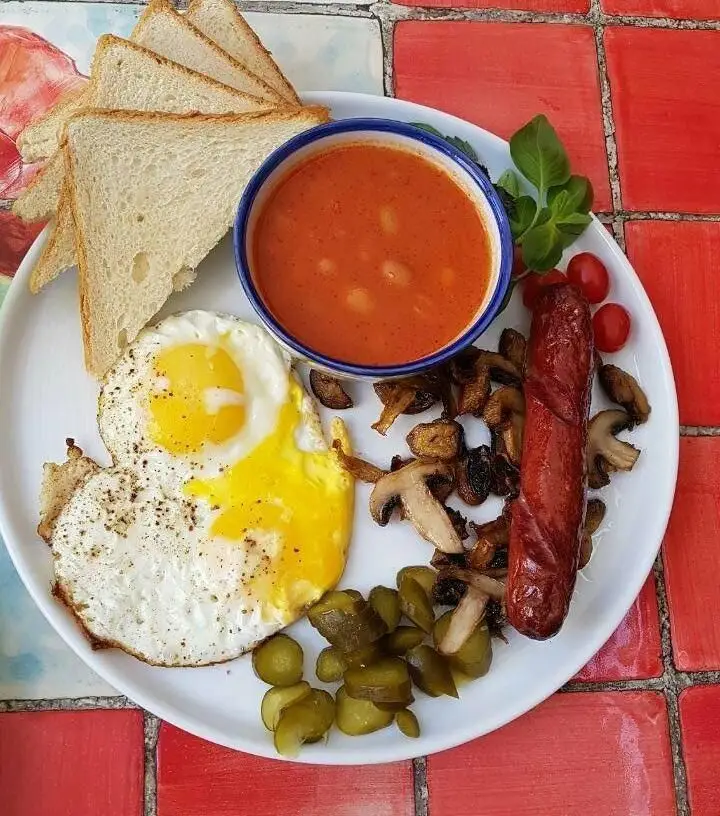 صبحانه کامل و لذیذ کافه مانا: شامل دو نیمرو، سوسیس، لوبیا، قارچ، نان تست و مخلفات تازه روی میزی با کاشی‌های سنتی.