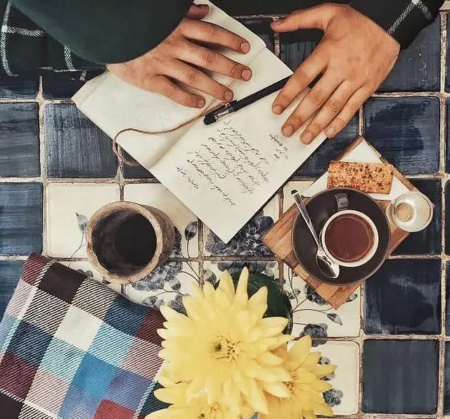 کافه مانا: صحنه‌ای دلنشین با دستانی مشغول نوشتن در دفترچه، فنجان قهوه گرم، گل‌های زرد و پتوی چهارخانه روی میز.