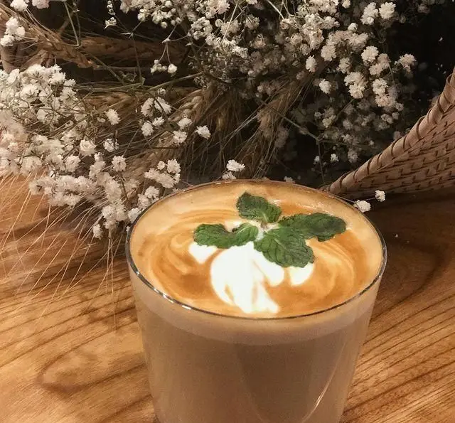 یک فنجان لاته گرم با طرح هنری شیر و برگ نعناع روی میز چوبی در کافه مانا، محیطی دلنشین برای تجربه قهوه‌ای باکیفیت و لحظاتی آرام. عطر تازه قهوه.