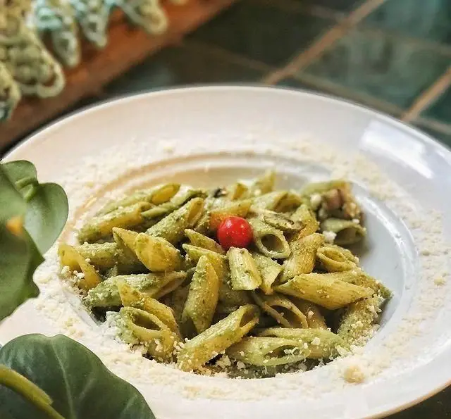 پاستا پنه با سس پستو سبز، تزئین شده با گوجه گیلاسی قرمز و پنیر رنده شده در یک کاسه سفید، غذایی لذیذ از منوی کافه مانا.