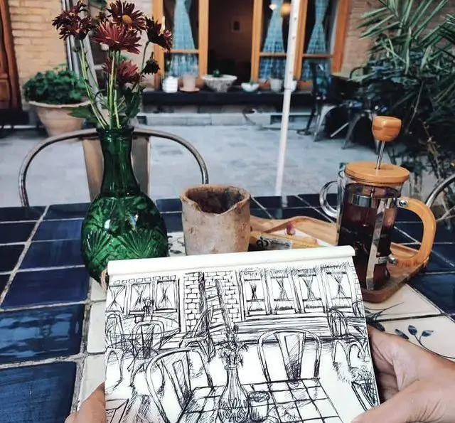 نمایی دلنشین از کافه مانا با میز کاشی‌کاری شده، گلدان گل و فرنچ‌پرس چای، به همراه دفترچه طراحی در دست. مناسب کافه گردی.