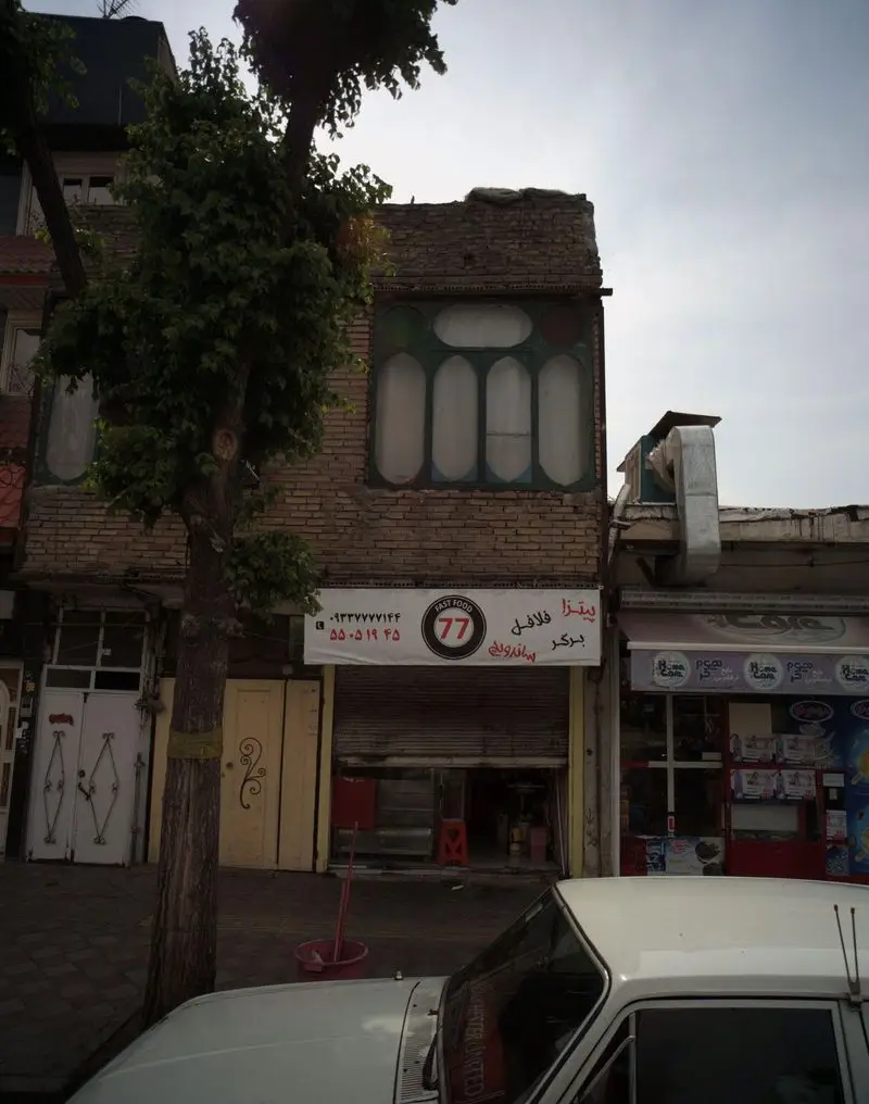 فست فود 77: تابلوی پیتزا، فلافل، برگر و ساندویچ روی ساختمان آجری، همراه با شماره تلفن. درخت و خودروی سفید جلوی مغازه.