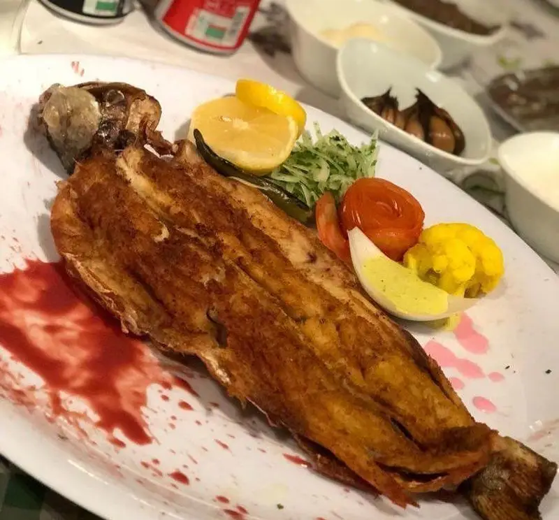 تصویری از ماهی کامل سرخ شده با پوستی طلایی، تزئین شده با لیمو، سبزیجات و گوجه، غذای لذیذ رستوران ماه و مهر.