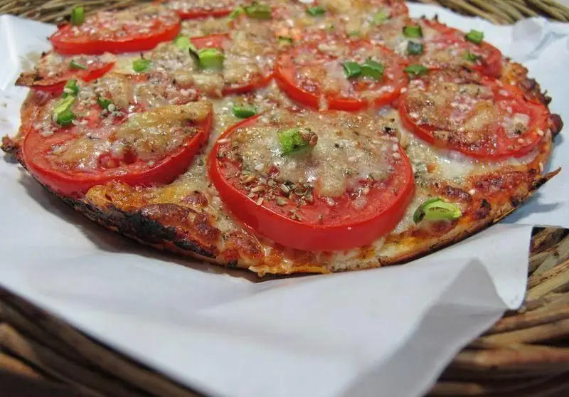 پیتزا آلونک: یک پیتزای گرد خوشمزه با گوجه‌فرنگی تازه، پنیر آب‌شده طلایی، چاشنی و پیازچه سبز. آماده سرو در سبد حصیری.