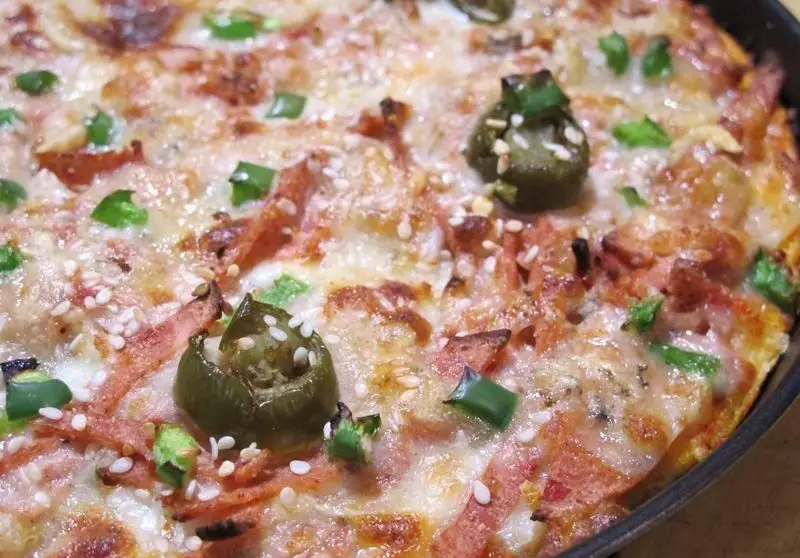 پیتزا آلونک: نمای نزدیک از پیتزای داغ و لذیذ با پنیر طلایی، کالباس یا ژامبون، فلفل هالاپینو و کنجد. ایده‌آل برای یک وعده خوشمزه.