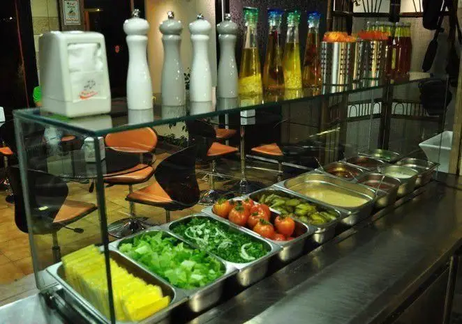 نمایی از سالاد بار سلف سرویس پیتزا آلونک با مخلفات تازه شامل کاهو، گوجه، خیارشور و انواع سس در محیطی دلنشین.