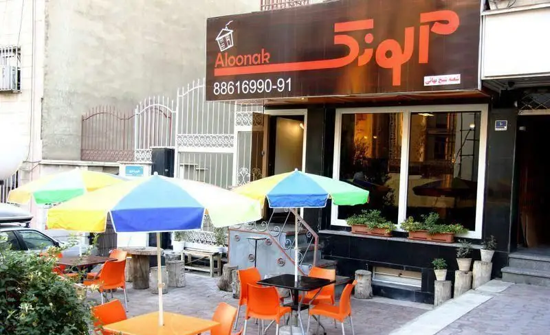 تصویر رستوران پیتزا آلونک (Aloonak) با تابلوی برند، فضای باز دلنشین، چترهای رنگارنگ و صندلی‌های نارنجی آماده پذیرایی.