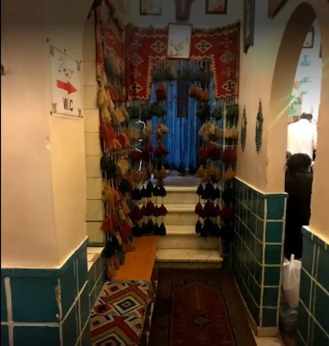 دکوراسیون داخلی سنتی و جذاب دیزی سرای 110. راهرویی با آویزهای رنگارنگ، قالیچه ایرانی و کاشی‌کاری فیروزه‌ای که فضایی دلنشین برای مشتریان ایجاد کرده است.