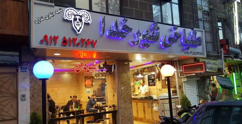 تصویر بیرونی طباخی شیر خدا با لوگو و تابلوی نئون، نمایش مشتریان داخل و ارائه بهترین کله پاچه و غذای سنتی ایرانی.