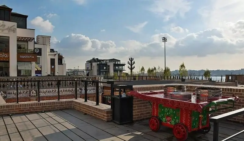 نمای بیرونی بام لند تهران با فروشگاه سوآچ، نرده‌های سنتی، چرخ دستی رنگارنگ و دریاچه چیتگر زیر آسمان آبی.