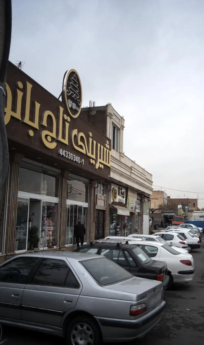 نمای بیرونی شیرینی‌فروشی تاج بانو با تابلوی طلایی، ورودی شیشه‌ای و شماره تماس ۴۴۳۳۰۳۴۰-۱ در خیابانی با خودروهای پارک‌شده.