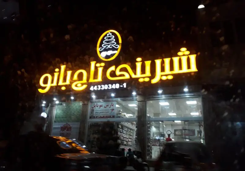 تصویر شب از تابلوی درخشان شیرینی فروشی تاج بانو با لوگوی منحصر به فرد و شماره تلفن ۴۴۳۳۰۳۴۰-۱، مکانی برای خرید انواع شیرینی و دسر.