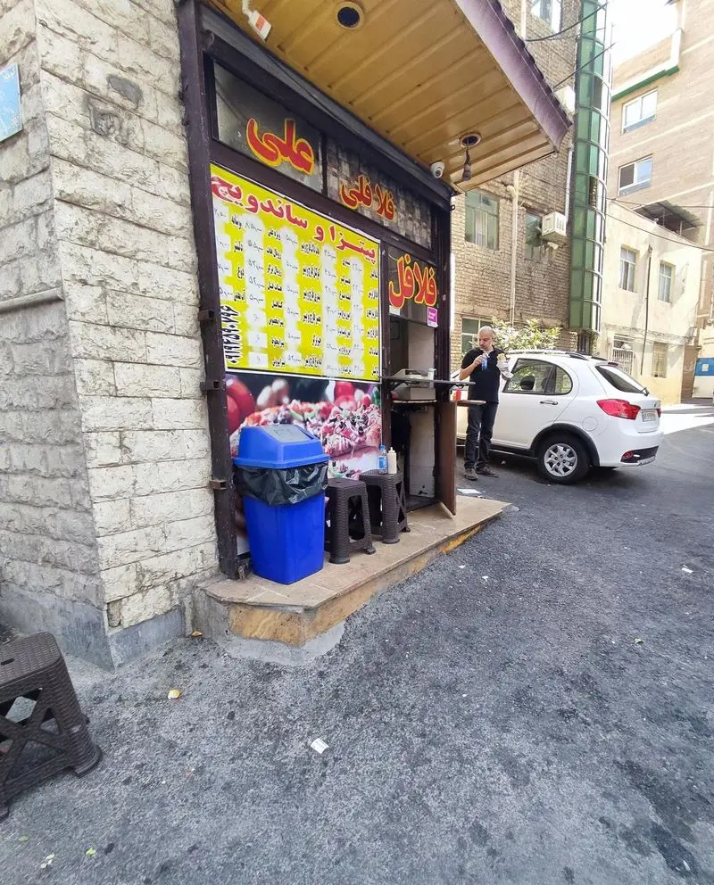 نمای بیرونی فلافلی علی با منوی پیتزا، ساندویچ و فلافل روی دیوار. مردی در حال خوردن غذا کنار ماشین پارک شده. یک مغازه اغذیه فروشی کوچک و فعال.