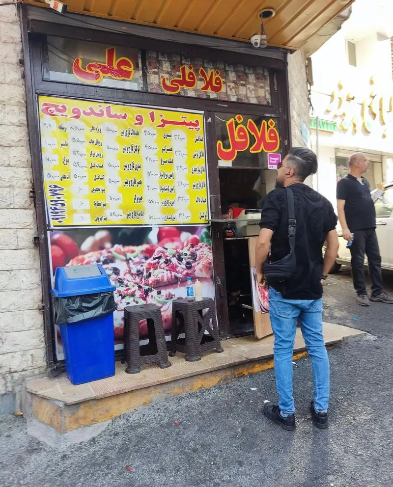 نمای بیرونی اغذیه فلافلی علی با تابلوهای قرمز "فلافلی علی" و "فلافل". منوی پیتزا و ساندویچ روی دیوار، و یک مشتری که منو را مشاهده می کند.