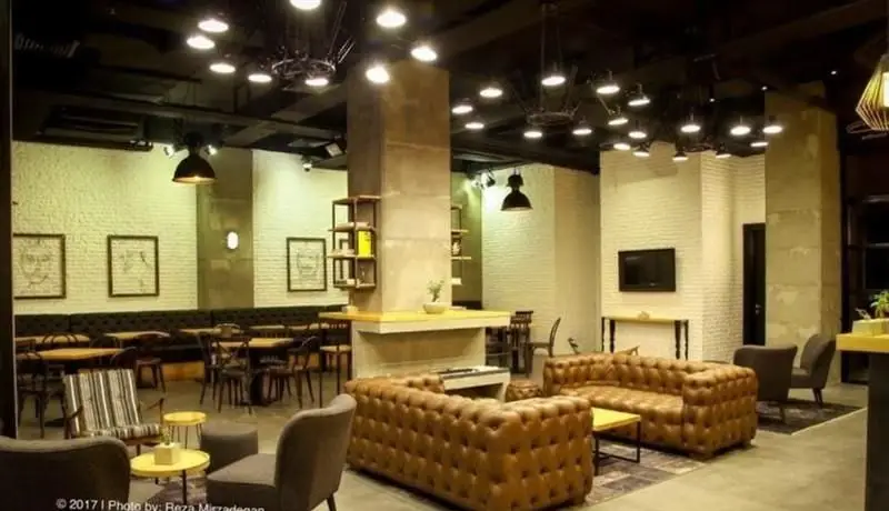 نمای داخلی کافی شاپ تیوان با طراحی مدرن-صنعتی، شامل مبلمان چرمی، میز و صندلی‌های متنوع، دیوارهای آجری و ستون‌های بتنی، فضایی دلنشین و کاربردی.