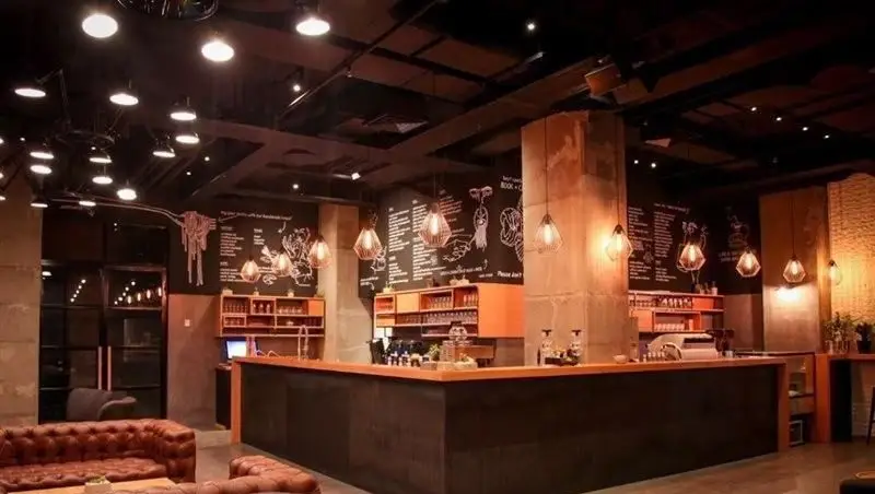 نمای داخلی جذاب کافی شاپ تیوان، فضایی مدرن و دنج با مبلمان چرمی، بار سرو قهوه، نورپردازی گرم و دیوار مشکی منو گچی.