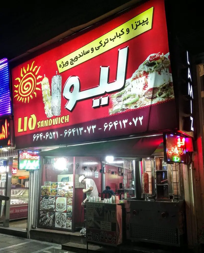 نمای شبانه فست فود لیو با تابلوی روشن، ارائه دهنده پیتزا، ساندویچ، کباب ترکی و شاورما. مشاهده آشپز در حال آماده سازی غذا.