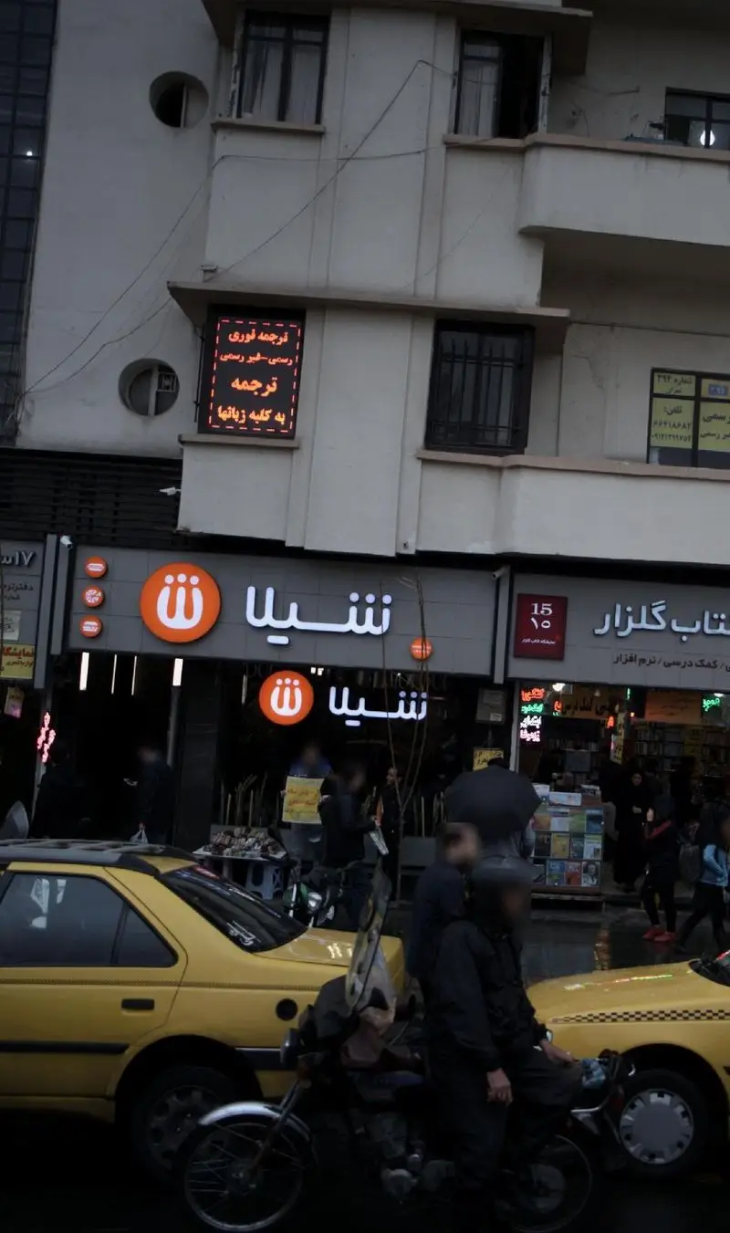 نمای بیرونی شعبه فست فود شیلا در تهران، با لوگوی نارنجی، در یک روز شلوغ شهری کنار کتابفروشی و دفتر ترجمه، همراه با تاکسی و موتور.