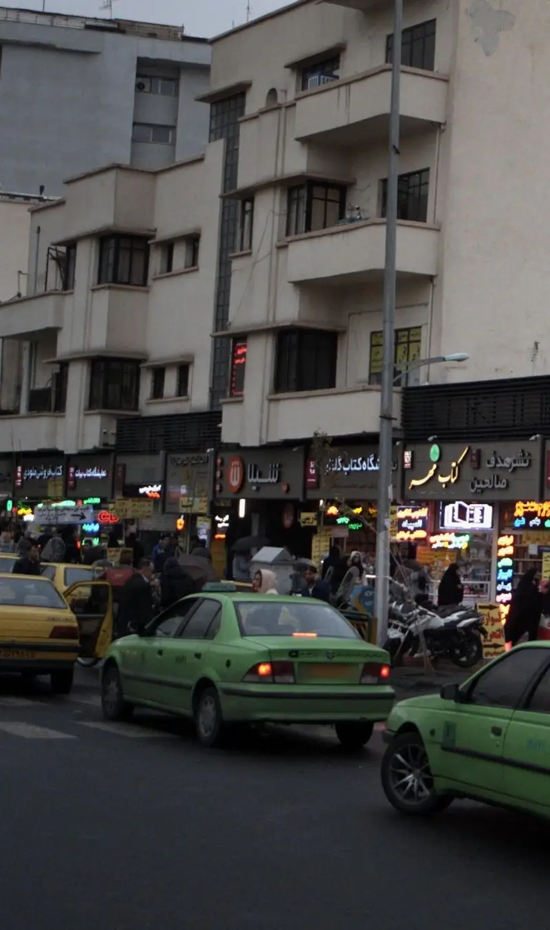 کسب و کار شیلا در یک خیابان شلوغ در ایران، با نمای ساختمان و تابلوهای فروشگاهی دیگر، همراه با تاکسی‌های سبز و مردم در رفت و آمد.