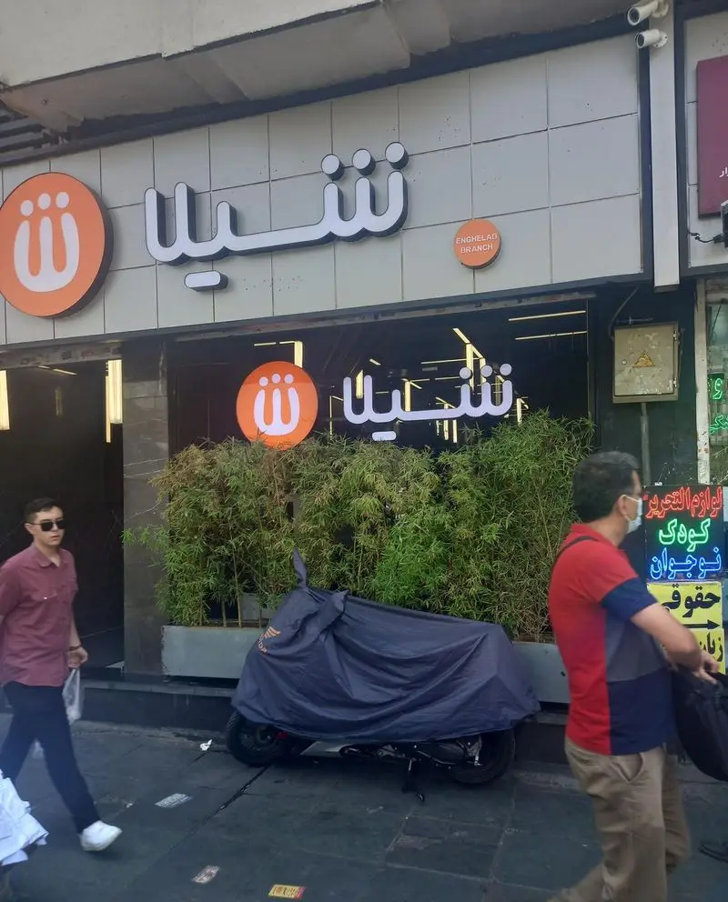 نمای بیرونی جذاب و مدرن شعبه انقلاب رستوران فست فود شیلا در تهران با لوگوی نارنجی، نوشته فارسی و حضور مردم در پیاده‌رو.