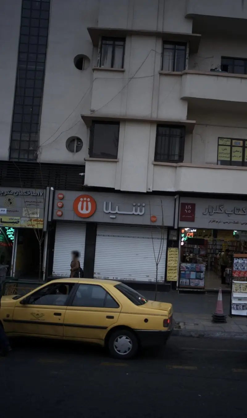 نمای بیرونی فروشگاه فست فود شیلا با لوگوی نارنجی مشخص، کرکره‌های بسته، و یک تاکسی زرد رنگ در خیابان شهر.