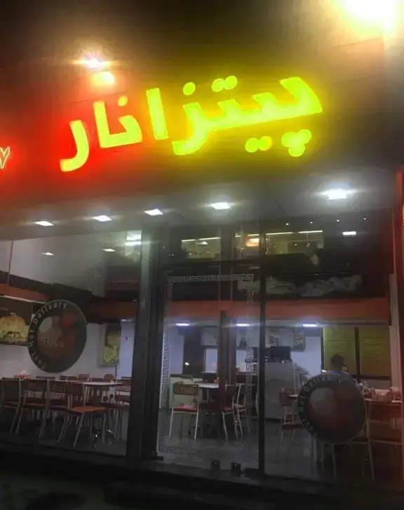 نمای شبانه فست فود نار با تابلوی نئونی درخشان پیتزانار و بازتاب نور، شامل ورودی و فضای داخلی رستوران با میز و صندلی.