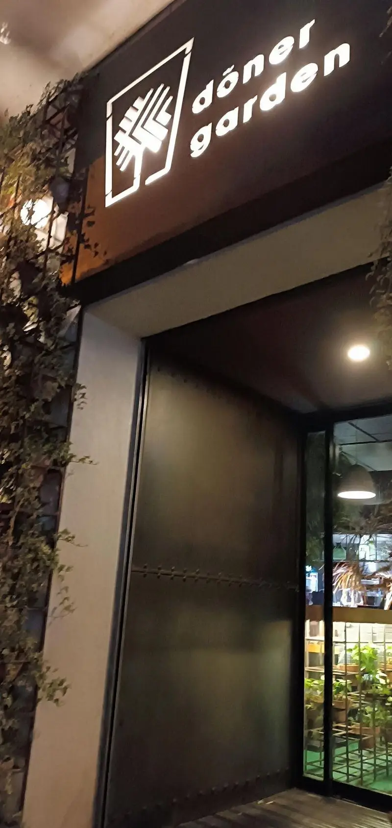 ورودی مدرن و تابلوی روشن فست فود دونر گاردن پاسداران در شب، با دیوارهای سبز و درب فلزی جذاب، آماده میزبانی.