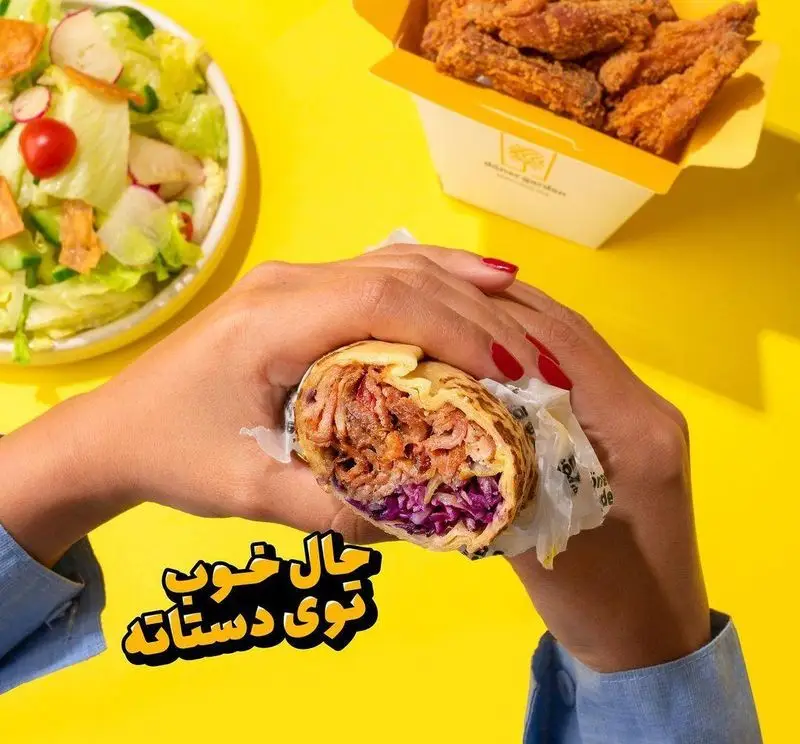 دستان فردی که ساندویچ دونر کباب تازه و خوشمزه از فست فود دونر گاردن پاسداران را در دست دارد، کنار سالاد و مرغ سوخاری.