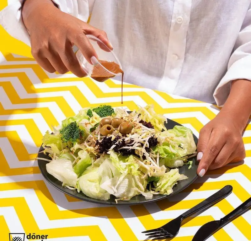 تصویری از دستانی که سس قهوه‌ای را روی سالاد تازه و رنگارنگ با کاهو، بروکلی و زیتون در فست فود دونر گاردن پاسداران می‌ریزند. غذای سالم و رژیمی.