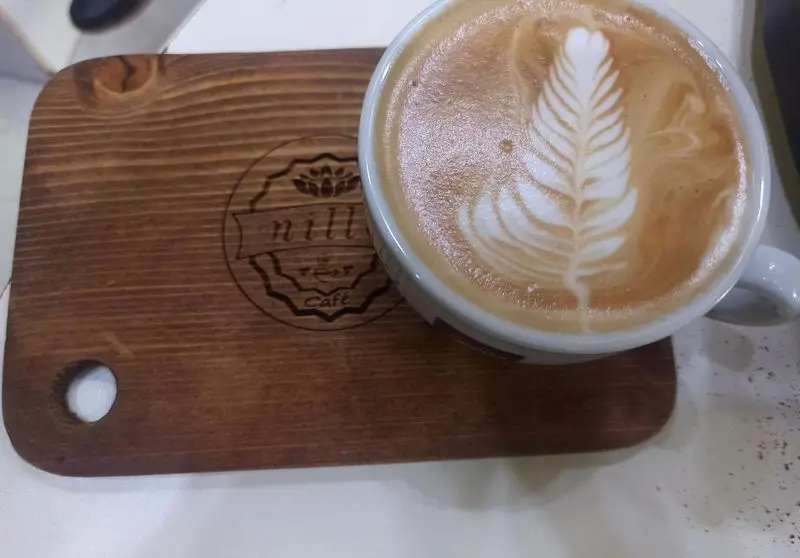 یک فنجان لاته آرت هنرمندانه با طرح برگ، روی تخته چوبی با لوگوی "nill Café" از کافه نیلو، برای علاقه‌مندان به قهوه و محیطی دلنشین