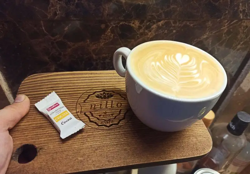 تصویری از یک لاته آرت دلنشین (قلب و برگ) در فنجان سفید، روی سینی چوبی برند کافه نیلو با لوگوی حک شده، به همراه ساشه کاکائو سالارگو.