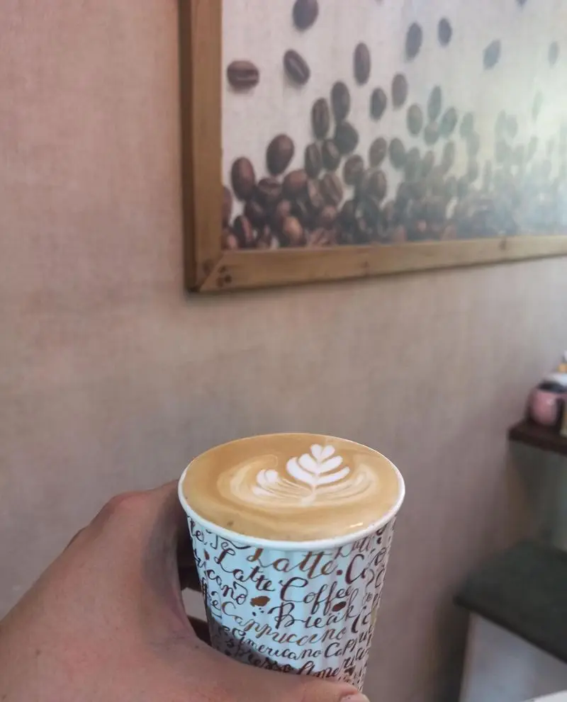 فنجان لاته آرت با طرح برگ، سرو شده در کافه نیلو. فنجان قهوه با نوشته‌های لاته و کاپوچینو، و تابلوی دانه قهوه در پس‌زمینه.