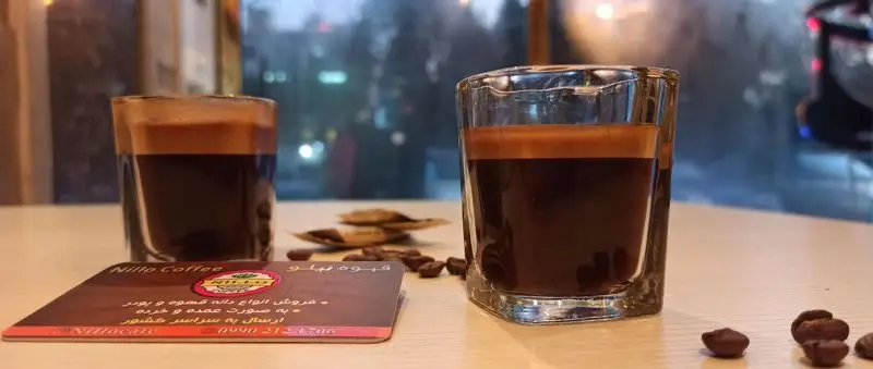 دو فنجان قهوه اسپرسو با کرمای طلایی، کارت ویزیت کافه نیلو و دانه های قهوه. فروش عمده و خرده انواع قهوه با ارسال به سراسر کشور.