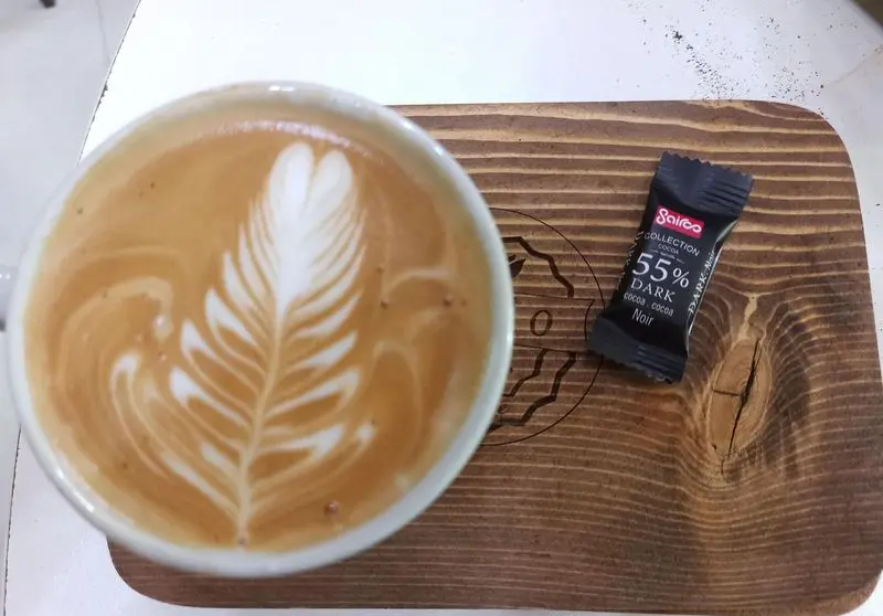 لاته آرت هنرمندانه با طرح برگ (پر) در کافه نیلو، همراه با شکلات تلخ ۵۵٪ Saice روی تخته سرو چوبی. تجربه‌ای دلنشین.