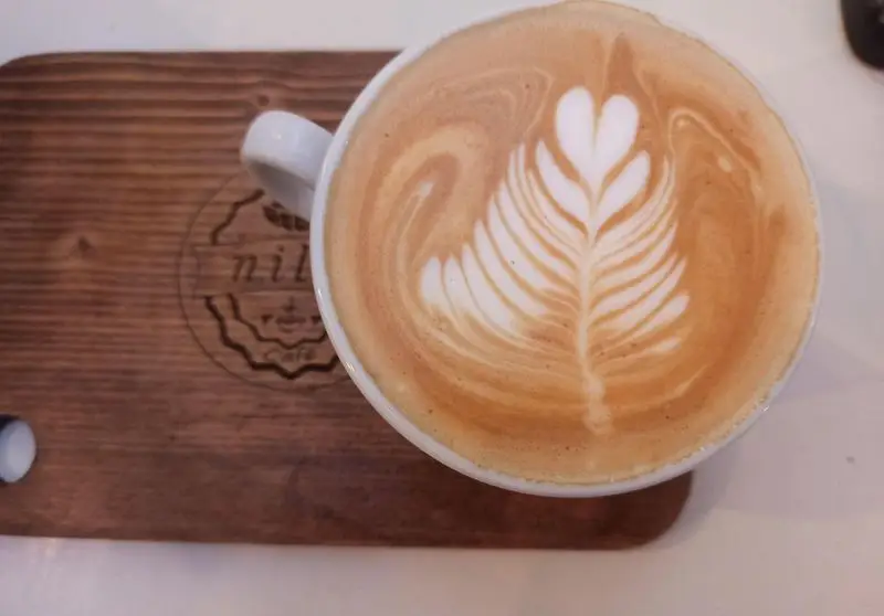 لاته آرت زیبا با طرح برگ و قلب در کافه نیلو، سرو شده بر روی تخته چوبی، نمادی از کیفیت و هنر قهوه‌سازی.