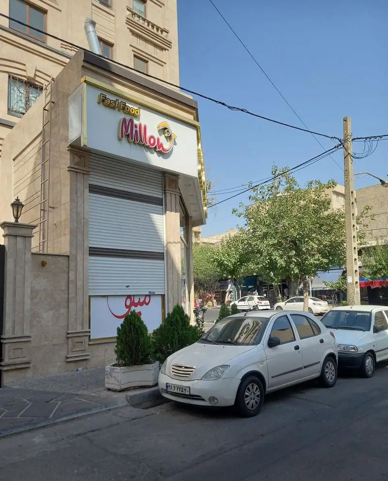 تصویری از نمای بیرونی فست فود میلو، با تابلوی درخشان "Fast Food Millow" و شخصیت زرد چشم، کرکره بسته و ماشین‌ها در روز آفتابی.