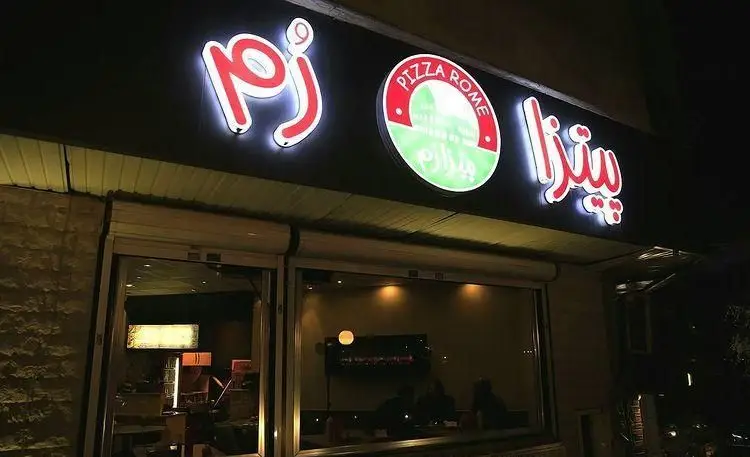 تصویری از تابلوی نورانی و چشم‌نواز "پیتزا رم" در شب، نمای بیرونی این رستوران فست فود با لوگوی جذاب و نام‌های فارسی و انگلیسی.