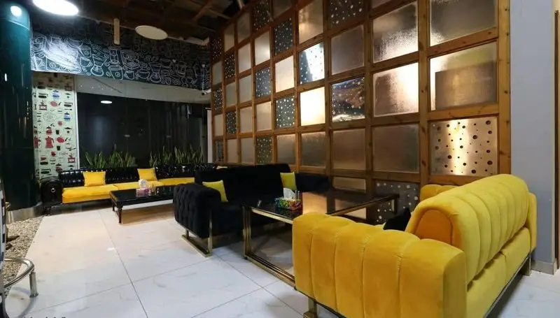 نمای داخلی کافه رستوران پلاس شیم با مبلمان مدرن و راحت زرد و مشکی، دیوار دکوراتیو چوبی و طرح‌های هنری قهوه، فضایی دعوت‌کننده.